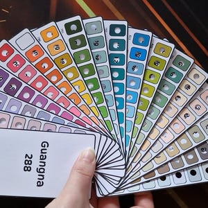 Puede incluir: Una mano sostiene un abanico de muestras de color, cada una mostrando un tono y un número únicos. Las muestras van desde rojos y naranjas vibrantes hasta azules y verdes fríos. También es visible una tarjeta blanca con el número "288" y el nombre "Guangna".