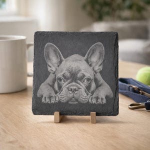 May include: Square slate coaster featuring an engraved image of a French Bulldog. The dog is depicted in a realistic style, resting its paws on the bottom edge. The coaster is displayed on a small wooden stand.