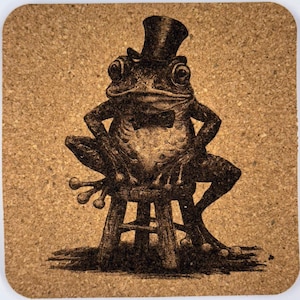May include: A square cork coaster featuring a detailed illustration of a frog wearing a top hat and bow tie, seated on a small stool. The frog has its arms crossed, and the design is in shades of brown.