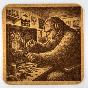 May include: Square cork coaster with a detailed illustration of a Bigfoot character wearing a headset and working on a computer. The image includes the text "I WANT TO BELIEVE" and alien-themed posters. The coaster has a brown color.