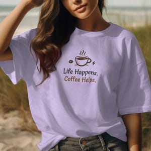 Peut inclure: T-shirt lavande clair avec l'inscription "Life Happens. Coffee Helps." et un motif sur le thème du café. Le motif comprend une tasse de café, un grain de café et un éclair. Le t-shirt est un vêtement décontracté de tous les jours.