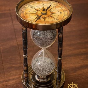 Könnte beinhalten: Eine antike Messing-Sanduhr mit Kompass. Der Kompass hat ein detailliertes Zifferblatt mit Richtungsmarkierungen. Die Sanduhr enthält grauen Sand und wird von einem dekorativen Ständer getragen. Auf dem Sockel steht der Text "ZIYA NAUTICAL".