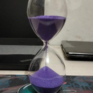 Könnte beinhalten: Eine Sanduhr aus klarem Glas mit abgerundeter Form, die leuchtend violetten Sand enthält. Der Sand fließt derzeit durch einen schmalen Hals vom oberen in den unteren Glaskolben und bildet einen kleinen kegelförmigen Haufen am Boden.