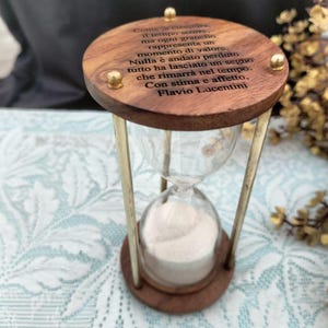 May include: A wooden hourglass with brass detailing and a glass chamber filled with white sand. The top wooden section features engraved text. The hourglass is a decorative item.