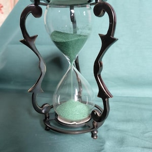 Könnte beinhalten: Eine Sanduhr mit einem dunklen Metallrahmen, geschwungenen, verzierten Stützen sowie runden Platten an Ober- und Unterseite. Die klaren Glaskolben enthalten feinen grünen Sand, der durch den schmalen Hals in der Mitte rieselt.