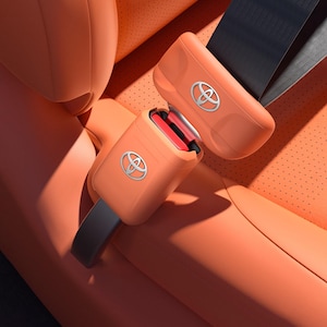 May include: An orange car seat belt buckle and latch mechanism featuring the silver Toyota logo on both components. The buckle is positioned above the receptacle, which is integrated into an orange perforated leather car seat surface.