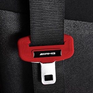 May include: Close-up of a black seatbelt with a red seatbelt buckle. The buckle has the text "AMG" in white on a black background. The seatbelt is fastened.
