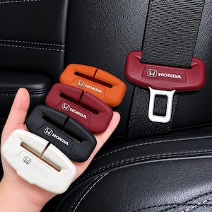 May include: A collection of Honda seat belt buckle covers in various colors: white, black, red, and orange. The covers have the Honda logo and text. One red cover is attached to a black seat belt.