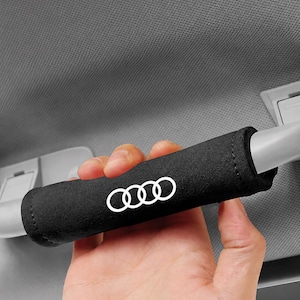 May include: Black car handle cover with the Audi logo in white. The cover is cylindrical and made of a soft, textured material. The Audi logo consists of four interlocking rings. The handle cover is designed to fit over a car's interior handle.