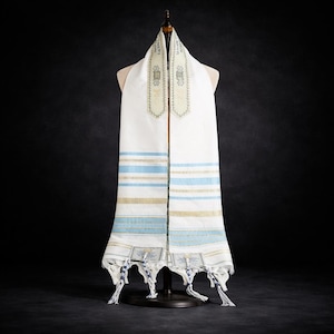 Peut inclure: Un tallit blanc, châle de prière juif, est présenté sur un mannequin. Le châle présente des rayures bleues et dorées, avec des broderies décoratives en haut et des glands en bas. Le fond est de couleur sombre.