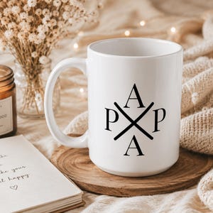 Pode incluir: Uma caneca de cerâmica branca com um design gráfico preto das letras "APA" em forma de cruz. A caneca está sobre um descanso de madeira, com uma vela acesa e um livro. O livro tem o texto "take your time to make your soul happy."