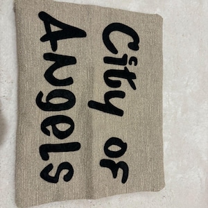 May include: A rectangular beige pillow with the words "City of Angels" embroidered in large, black, cursive letters. The pillow has a textured surface.
