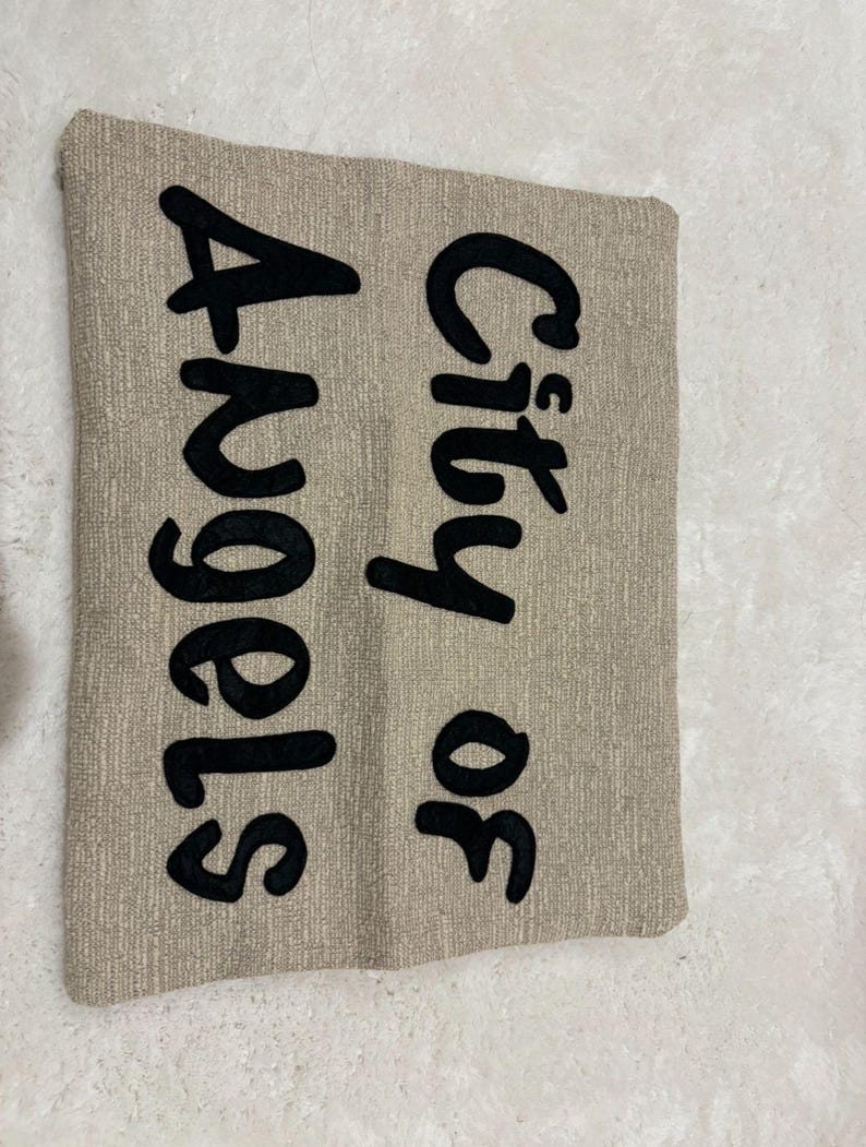 May include: A rectangular beige pillow with the words "City of Angels" embroidered in large, black, cursive letters. The pillow has a textured surface.