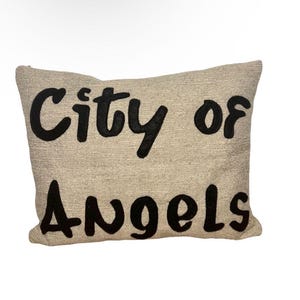 Op de afbeelding: Een rechthoekig beige kussen met de woorden "City of Angels" geborduurd in grote, zwarte, gestileerde letters. Het kussen heeft een textuur, wat wijst op linnen of een vergelijkbare stof.