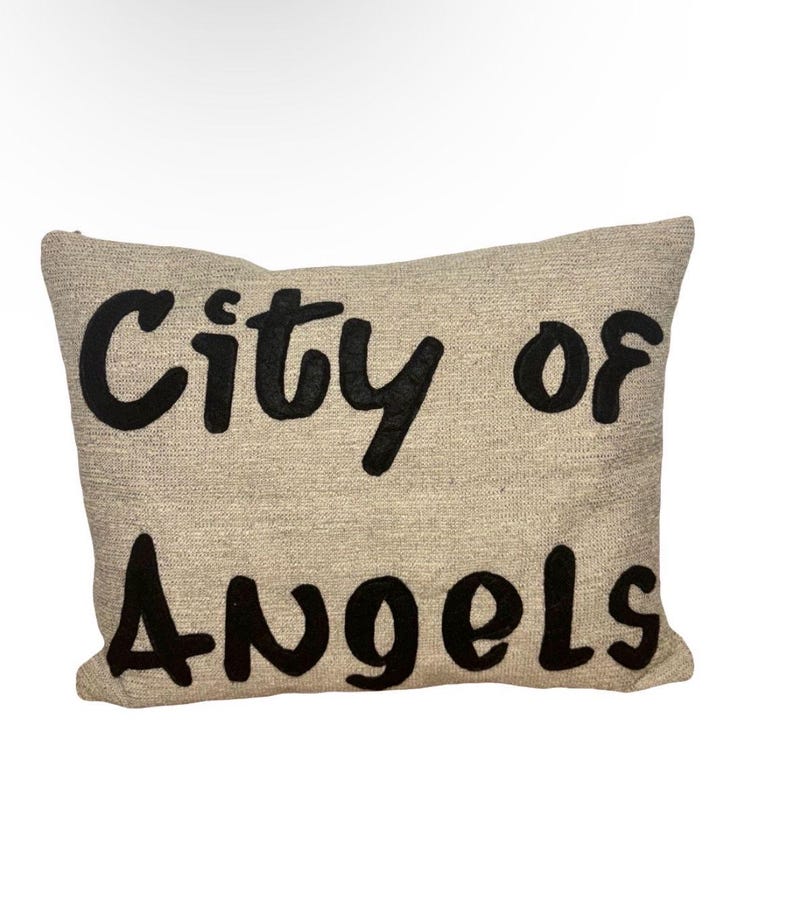 May include: A rectangular beige pillow with the words "City of Angels" embroidered in large, black, stylized letters. The pillow has a textured appearance, suggesting a linen or similar fabric.