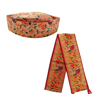 May include: A tan-colored cap with a structured band and a rounded top, featuring a vibrant floral pattern in red, orange, green, and purple. Beside it is a long, rectangular sash with matching floral motifs and a bold red border with tassels.