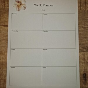 May include: A white week planner with a floral design in the upper left corner. The planner is divided into sections for each day of the week, with space for notes. The text "Week Planner" is at the top.