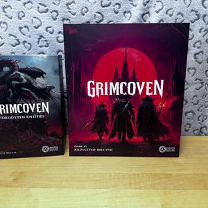 Può includere: Due scatole di giochi da tavolo con il titolo "Grimcoven" in lettere bianche. La scatola più grande presenta uno sfondo rosso con figure silhouette. La scatola più piccola ha uno sfondo scuro con un'illustrazione di mostri. È visibile anche il testo "Forgotten Entities".
