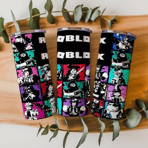 May include: Three black insulated tumblers with a colorful Roblox design. Each tumbler features a black background with the word "ROBLOX" in white and a grid of Roblox character illustrations in various colors. The tumblers are displayed on a wooden surface.