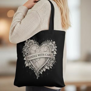 Könnte beinhalten: Schwarze Tragetasche mit einem Herzdesign, das von Rosen und Dornen umgeben ist. Das Herz ist zerbrochen und trägt ein Banner mit der Aufschrift "HANDLE WITH CARE". Das Design ist in Grau- und Weißtönen gehalten.