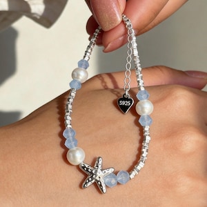 May include: A silver-toned bracelet featuring a central starfish charm, white pearls, and light blue faceted beads. The bracelet includes a silver chain with a small heart-shaped tag engraved with S925 and an adjustable extender chain.