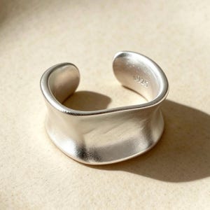 May include: A wide, open-style silver ring with a smooth, matte finish and a curved, undulating band. The interior of the ring is clearly stamped with the text S925, indicating the metal composition of this solid, polished silver jewellery piece.