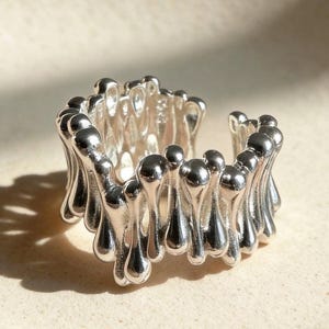 May include: A silver ring with an unusual, organic design. The ring has a series of rounded, droplet-shaped elements that encircle the band. The open design allows for an adjustable fit. The ring is made of a shiny, reflective metal.