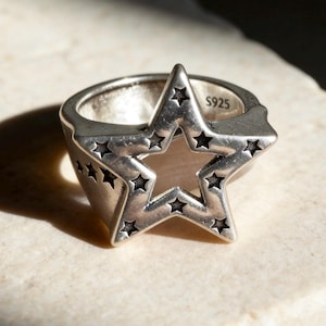 May include: A silver-toned ring featuring a prominent star-shaped cutout design on the face. The surface of the star is decorated with smaller engraved star motifs. The interior of the band is clearly stamped with the text S925, indicating the metal.