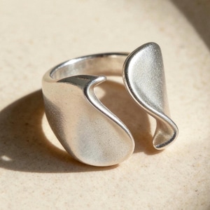 May include: A silver ring with an unusual, open design. The ring has two curved, petal-like forms that encircle the finger. The metal has a matte finish, and the ring is set against a neutral background.