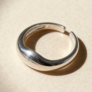 May include: A silver ring with a smooth, rounded design. The ring has a thick band that tapers slightly towards the open end. The ring is made of a shiny, reflective metal, and is likely a fashion accessory.