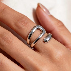 May include: A polished silver ring featuring an open, bypass-style design. The band consists of a smooth, rounded metal loop that curves around the finger, ending in a separate, rounded silver bead element that sits adjacent to the main loop.