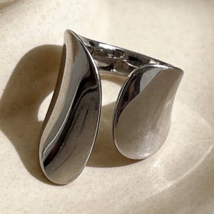 May include: A silver-toned ring with a unique, open design. The ring features two curved, teardrop-shaped elements that wrap around the finger. The polished metal reflects light, creating highlights and shadows. The ring is displayed on a neutral-toned surface.