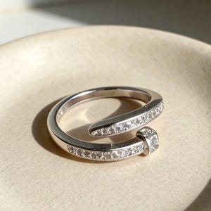 May include: A silver-toned bypass ring featuring a wrap-around nail design. The band is encrusted with a row of small, clear, sparkling stones along the outer surface, terminating in a flat, square-shaped head also set with a clear stone.
