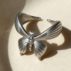 May include: A silver butterfly ring. The ring has a detailed butterfly design with open wings and a heart-shaped detail on the body. The ring is adjustable and made of a metallic material.