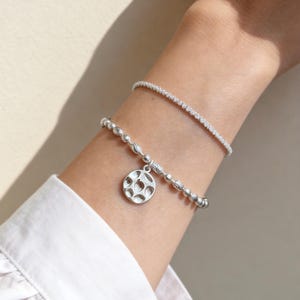 Peut inclure: Deux bracelets argentés portés ensemble. Le bracelet supérieur est une chaîne fine et délicate avec une finition texturée et scintillante. Le bracelet inférieur se compose de perles rondes lisses et d'une breloque circulaire à motif de six points.