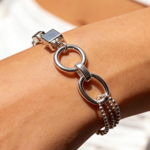 May include: A silver-toned bracelet featuring a polished square link, a circular ring, an oval link, and three delicate beaded chains that drape downwards. The metal components have a smooth, reflective finish and are connected in a linear sequence.