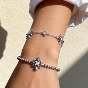 Peut inclure: Deux bracelets argentés. Le bracelet supérieur présente une chaîne délicate avec des breloques en forme de fleur alternées et de petites pierres claires. Le bracelet inférieur se compose d'une chaîne perlée avec une breloque florale centrale.