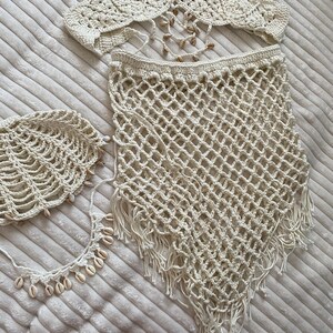 May include: Cream-coloured crochet bikini set, including a halter top, skirt, and headpiece. The halter top and headpiece are decorated with seashells. The skirt features a net-like pattern with fringe. The set is displayed on a textured, cream-coloured surface.