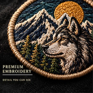 May include: A circular embroidered patch featuring a wolf head in profile, snow-capped mountains, a golden sun, and green pine trees. The border is stitched in tan thread. Text on the image reads: PREMIUM EMBROIDERY DETAIL YOU CAN SEE.