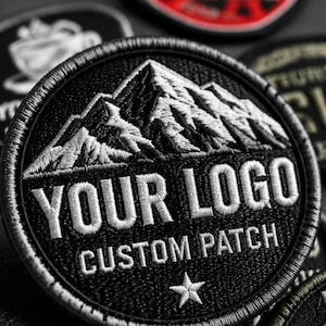 May include: A circular embroidered patch featuring a black background with a white mountain range graphic at the top. The text YOUR LOGO is embroidered in white, followed by the text CUSTOM PATCH and a small white star at the bottom edge.