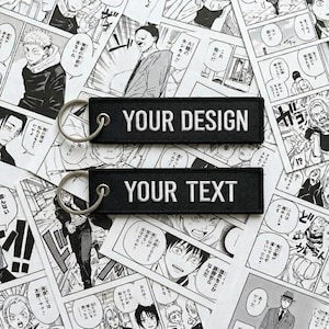 Könnte beinhalten: Zwei rechteckige Schlüsselanhänger aus schwarzem Stoff mit weißem Sticktext. Der obere Anhänger zeigt den Schriftzug YOUR DESIGN, der untere den Schriftzug YOUR TEXT. Jeder Anhänger ist mit einem silberfarbenen Metallring versehen.