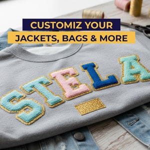 May include: A grey sweatshirt with the name "STELLA" appliqu&eacute;d in blue, pink, and teal letters with gold glitter trim. The text "CUSTOMIZE YOUR JACKETS, BAGS & MORE" is displayed in a banner above the name.