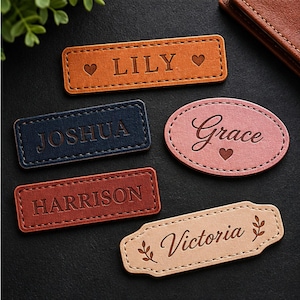 May include: Five rectangular and oval leather labels with stitched edges. The labels feature engraved names: LILY with hearts, JOSHUA, Grace with a heart, HARRISON, and Victoria with leaf accents. The colors include tan, dark blue, pink, and brown.