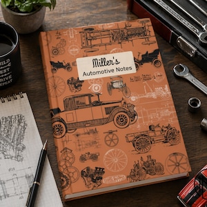 May include: An orange notebook titled "Miller's Automotive Notes" with vintage car illustrations. A black mug with "Build, Test, Drive, Repeat" text, a notepad with a V8 engine diagram, and a red toy car are also visible.