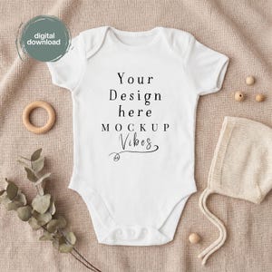 Könnte beinhalten: Ein weißer kurzärmeliger Babybody mit dem schwarzen Aufdruck Your Design here MOCKUP Vibes. Das Kleidungsstück wird zusammen mit einem Holzring, einer Strickhaube und einem Zweig aus getrocknetem Eukalyptus auf strukturiertem Stoff gezeigt.