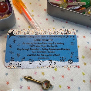 May include: A collection of small trinkets, including a key, dice, and a green toy car, arranged on a star-patterned surface. The image also features a business card with text and a pen. The items are in small metal tins.
