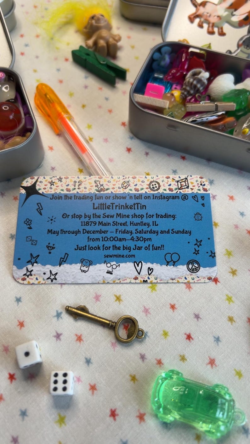 May include: A collection of small trinkets, including a key, dice, and a green toy car, arranged on a star-patterned surface. The image also features a business card with text and a pen. The items are in small metal tins.