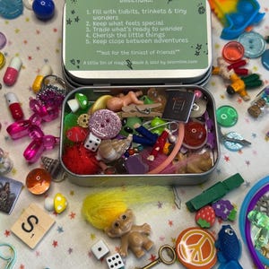 May include: A small, open metal tin filled with various trinkets, including a troll doll, dice, buttons, and miniature figurines. The tin is surrounded by other small items like buttons, beads, and a peace sign. The tin has text that reads "Fill with tidbits, trinkets & tiny wonders."