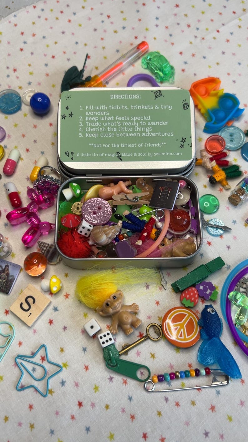 May include: A small, open metal tin filled with various trinkets, including a troll doll, dice, buttons, and miniature figurines. The tin is surrounded by other small items like buttons, beads, and a peace sign. The tin has text that reads "Fill with tidbits, trinkets & tiny wonders."