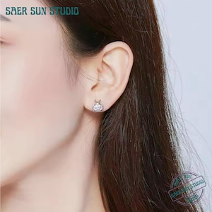 May include: A close-up of a person's ear with a small, round stud earring. The earring features a central, clear gemstone surrounded by a decorative setting. The text "SAER SUN STUDIO" is visible at the top of the image.